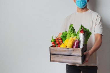 Koruyucu maskeli genç teslimatçı elinde tahta kutu tutuyor. Alışveriş kutusu olan bir kurye. Süpermarketten eve güvenli teslimat. COVID-19 koronavirüs koruması