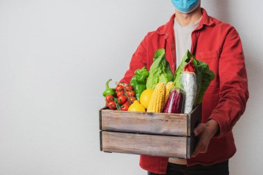 Koruyucu maskeli genç teslimatçı elinde tahta kutu tutuyor. Alışveriş kutusu olan bir kurye. Süpermarketten eve güvenli teslimat. COVID-19 koronavirüs koruması.