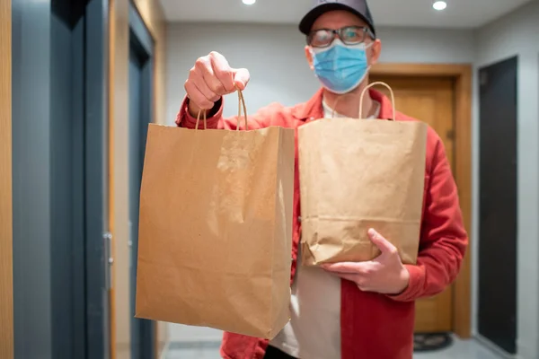 Delivery man in protective mask holding paper bag with food in the entrance. The courier gives the box with fresh vegetables and fruits to the customer.