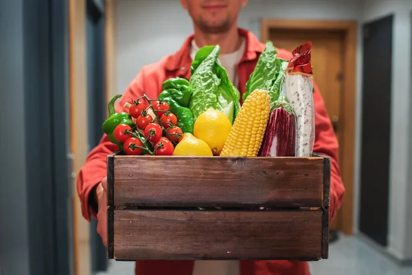 Delivery man holding paper bag with food in the entrance. The courier gives the box with fresh vegetables and fruits to the customer.