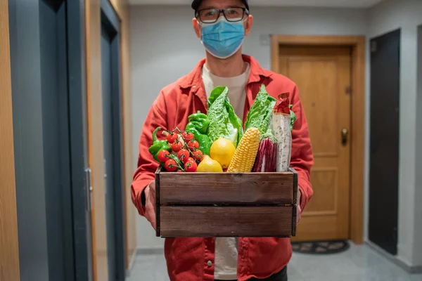 Delivery man in protective mask holding paper bag with food in the entrance. The courier gives the box with fresh vegetables and fruits to the customer.