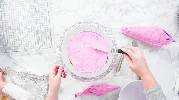 Flat lay. Step by step. Frosting round funfetti cake layers with pink buttercream frosting.