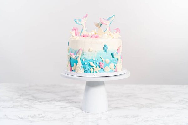 Mermaid-themed 3 layer vanilla cake decorated with chocolate mermaid tails and seashells on a white cake stand.
