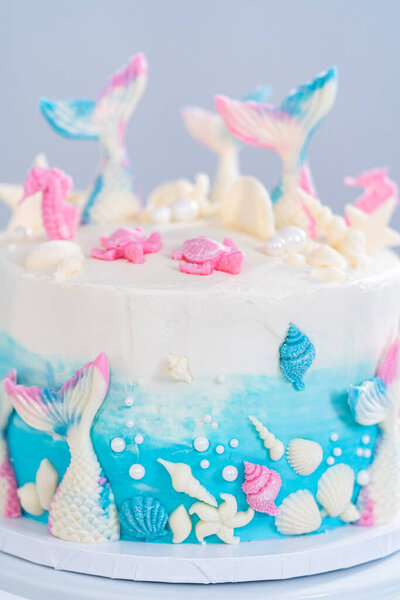 Mermaid-themed 3 layer vanilla cake decorated with chocolate mermaid tails and seashells on a white cake stand.