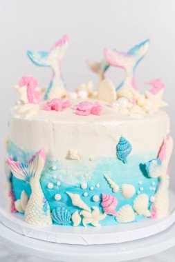 Deniz kızı temalı 3 katlı vanilyalı pasta çikolatalı deniz kızı kuyruğu ve deniz kabuğu ile süslenmiş beyaz pasta standı.