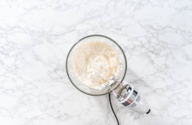 Flat lay. Whisking buttercream frosting in a glass mixing bowl with an electric hand mixer to prepare the peppermint buttercream frosting.