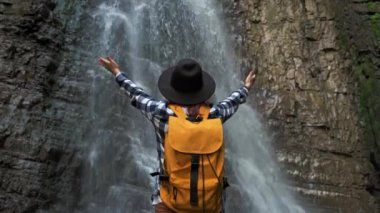 Şelalenin önünde duran sarı bir sırt çantası ve şapka takan bir gezgin kız. Yaşam Tarzı Seyahat Konsepti.