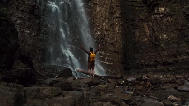 Traveler man with a yellow backpack standing on the background of a waterfall. — Stock Video