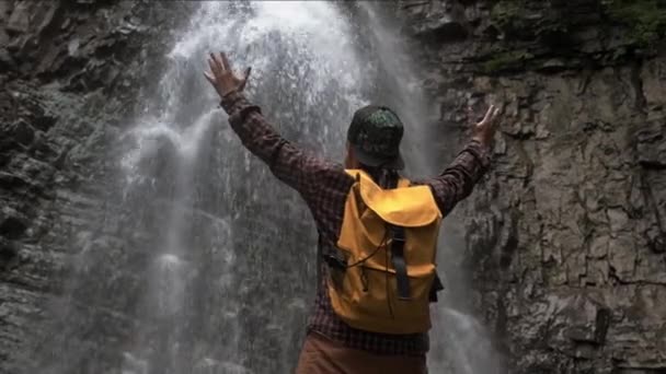 Resenär man med en gul ryggsäck står på bakgrunden av ett vattenfall. — Stockvideo