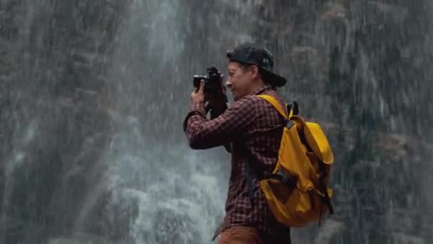Porträtt av en resenär fotograf med en gul ryggsäck står på bakgrunden av ett vattenfall gör ett foto — Stockvideo