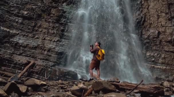 Travel man with a yellow backpack standing on the background of a waterfall makes a photo landscape. Travel lifestyle concept. Copy, empty space for text — Stock Video