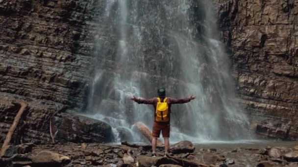 Viaggiatore uomo con uno zaino giallo in piedi sullo sfondo di una cascata. — Video Stock