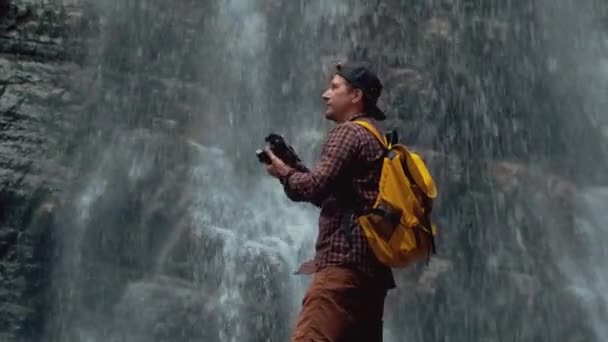 Retrato de um fotógrafo viajante com uma mochila amarela em pé no fundo de uma cachoeira faz uma foto — Vídeo de Stock