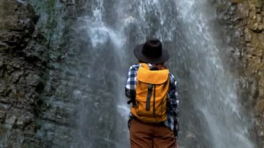 Şelalenin önünde duran sarı bir sırt çantası ve şapka takan bir gezgin kız. Yaşam Tarzı Seyahat Konsepti.