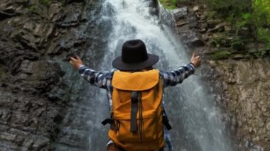 Şelalenin önünde duran sarı bir sırt çantası ve şapka takan bir gezgin kız. Yaşam Tarzı Seyahat Konsepti.