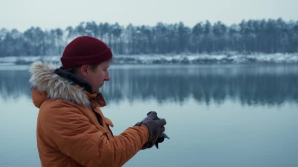 Ταξιδιώτης με πορτοκαλί μπουφάν φωτογραφίζει με μια σύγχρονη κάμερα στο φόντο ενός χειμερινού τοπίου — Αρχείο Βίντεο