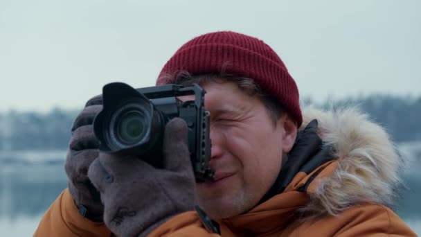 Travel man in an orange jacket and a red hat takes a picture with a modern camera against the background of a winter landscape. — Vídeo de Stock