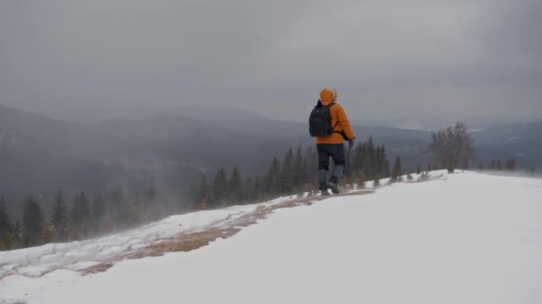 Hombre caminando a lo largo del camino en la cima de la colina cubierta de nieve. El hombre con una chaqueta amarilla camina en invierno caminando. vista desde atrás — Vídeos de Stock