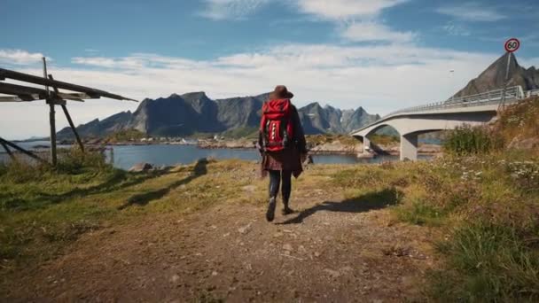 ノルウェーの小さな漁村の桟橋に赤いバックパックと帽子を持つヒップスター旅行者の女の子 ノルウェーの小さな漁村でロフトテンの美しい山々を背景に旅行者 — ストック動画