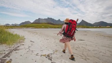 Büyük kırmızı sırt çantalı ve şapkalı bir turist kız dağlarda sahil boyunca yürüyor. Güzel Norveç yaz manzarası