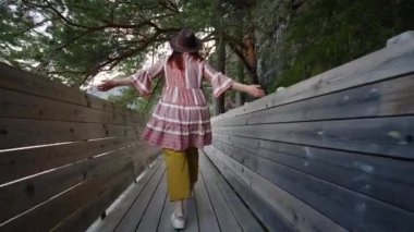 Şapkalı bir kadın, elleri novejede tahta bir köprüde yürür..