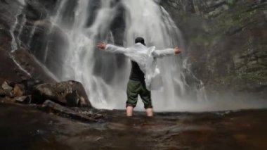 Şeffaf yağmurluk giyen mutlu adam Norveç 'te bir şelalenin dibinde elleri havada bir kayanın yanında duruyor..