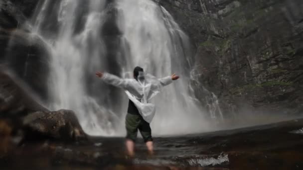 透明なレインコートを着た幸せな男が岩の近くのノルウェーの滝のふもとに手を上げて立っている — ストック動画
