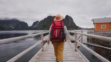 Gezgin ahşap iskeleye gider. Süveter, örgü şapka ve kırmızı bir sırt çantası giymişti. Arkadan bak. Lofoten Adaları Norveç.