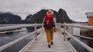 Gezgin ahşap iskeleye gider. Süveter, örgü şapka ve kırmızı bir sırt çantası giymişti. Arkadan bak. Lofoten Adaları Norveç.