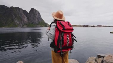 Gezgin ahşap iskeleye gider. Süveter, örgü şapka ve kırmızı bir sırt çantası giymişti. Arkadan bak. Lofoten Adaları Norveç.