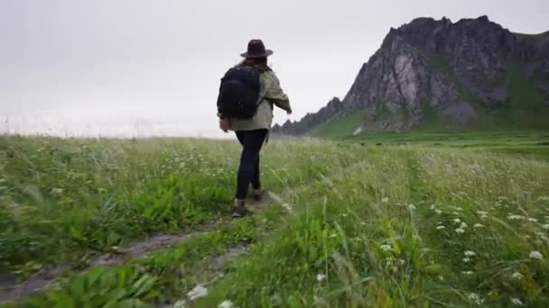 帽子をかぶった旅行者の女の子緑のジャケットとバックパック彼女は北欧の海の海岸に沿って歩く — ストック動画