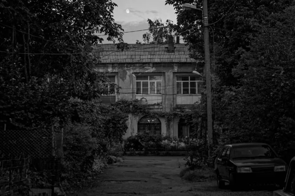 Czarno-biały odkryty fotografia małego miasteczka stary dwupiętrowy dom dziedziniec utonął w zieleni z starym samochodem zaparkowanym po stronie drogi w zmierzchu z księżycem nad chmurami w ciemnym niebie — Zdjęcie stockowe