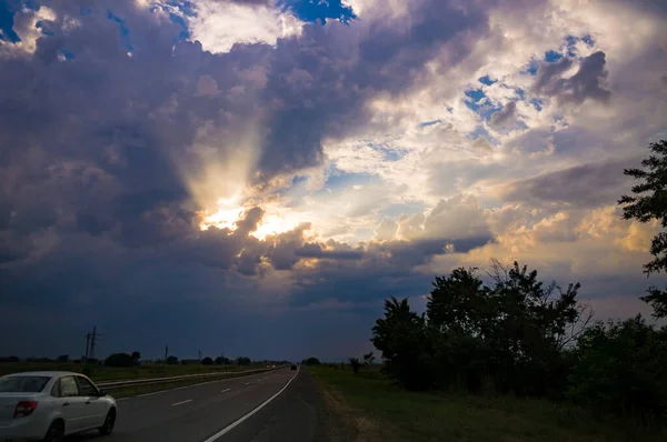 Πολύχρωμο φθινόπωρο βράδυ τοπίο με ακτίνες του ήλιου ακτινοβολούν από ένα άνοιγμα στο δραματικό συννεφιασμένο ουρανό ηλιοβασιλέματος πάνω από την εθνική οδό με ένα λευκό αυτοκίνητο — Φωτογραφία Αρχείου
