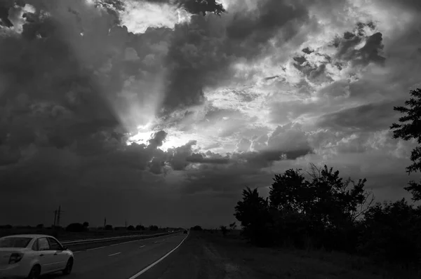 Ασπρόμαυρη εικόνα του φθινοπώρου τοπίου βράδυ με ακτίνες του ήλιου ακτινοβολούν από ένα άνοιγμα στο δραματικό συννεφιασμένο ουρανό ηλιοβασιλέματος πάνω από την εθνική οδό με ένα λευκό αυτοκίνητο — Φωτογραφία Αρχείου