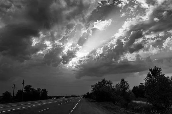 Ασπρόμαυρη εικόνα του φθινοπώρου τοπίου βράδυ με ακτίνες του ήλιου ακτινοβολούν από ένα άνοιγμα στο δραματικό συννεφιασμένο ουρανό ηλιοβασιλέματος πάνω από την εθνική οδό — Φωτογραφία Αρχείου