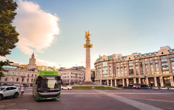 Tbilisi, Georgia - 07 23 2022: Green city bus coming from a roundabout in front of gilded statue of St. George atop a granite column on monumental Freedom Square in Downtown Tbilisi seen at summer