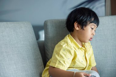 Sarı tişörtlü, siyah saçlı Asyalı çocuk tabletin üzerinde oturuyor. Pencerenin yanındaki oturma odasındaki gri koltuğun tadını çıkarıyorum..