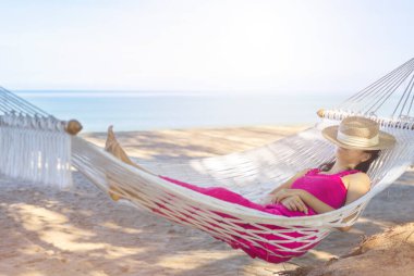 Asian woman relaxing in the hammock on tropical beach, njoy her freedom and fresh air, wearing stylish hat and clothes. Happy smiling tourist in tropics in travel vacation.