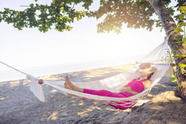 Asian woman relaxing in the hammock on tropical beach, njoy her freedom and fresh air, wearing stylish hat and clothes. Happy smiling tourist in tropics in travel vacation.