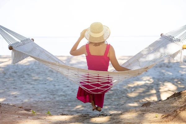 Tropikal plajda hamakta dinlenen Asyalı kadın, özgürlüğünün ve temiz havasının keyfini çıkarıyor, şık şapka ve giysiler giyiyor. Seyahat tatilinde tropiklerde gülümseyen mutlu turistler..