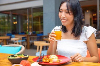 Kahvaltı eden bir kadın. Yumurtalı Benedict, karpuz, papaya, kavun, çarkıfelek meyvesi, portakal suyu ve kahve gibi meyveler. gri bir paspasa yerleştirilmiş..