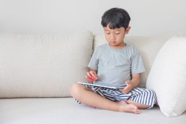 Gri tişörtlü, mavi çizgili şortlu sevimli mutlu Asyalı çocuk krem rengi kanepesinde tabletiyle oynarken eğleniyor. Mobil ekrana bakıyorum..