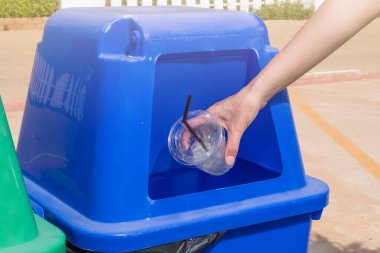 Yakından bakınca mavi geri dönüşüm kutusuna boş plastik bardak atıyor. Geri dönüşüm merkezine götürülmeden önce geri dönüşümleri tutmak için kullanılan bir konteynır.