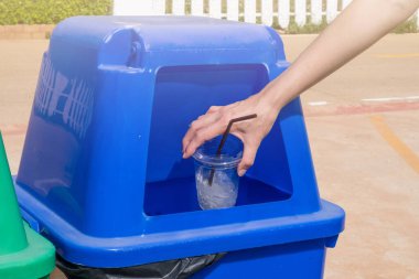 Yakından bakınca mavi geri dönüşüm kutusuna boş plastik bardak atıyor. Geri dönüşüm merkezine götürülmeden önce geri dönüşümleri tutmak için kullanılan bir konteynır.