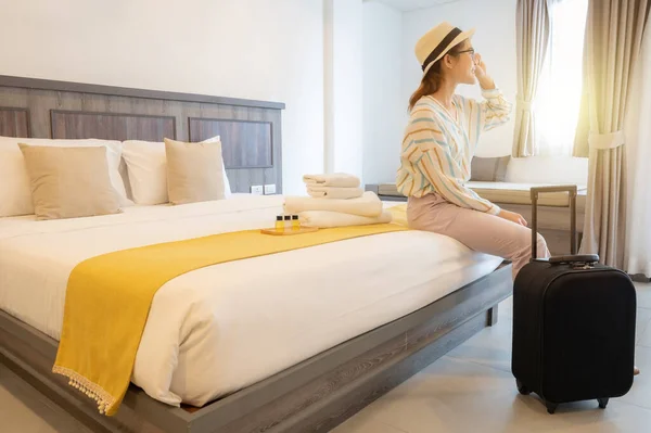 Tourist woman sitting on bed and looking to beautiful view outside the room with her luggage in hotel bedroom after check-in. Conceptual of travel and accommodation.