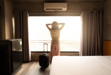 Pencere kenarında duran turist kadının, otele giriş yaptıktan sonra bavuluyla güzel bir manzara izlemesi. Seyahat ve tatil kavramı.