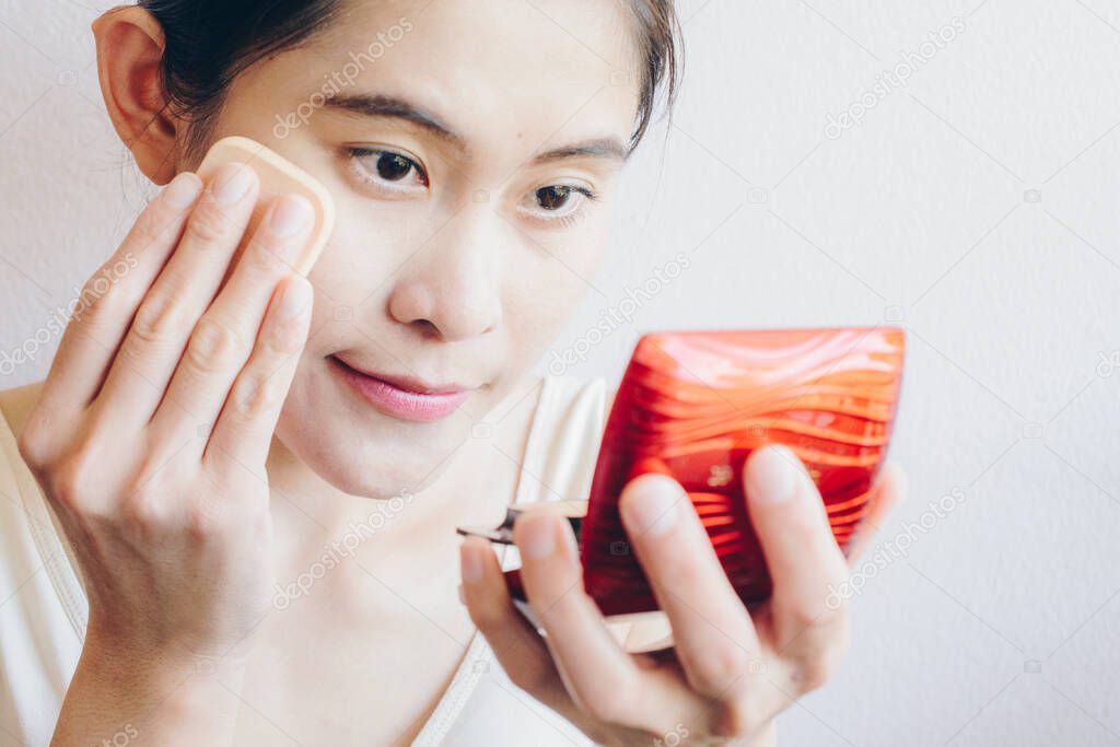Portrait of Asian woman applying foundation powder puff on her face to cover flaws and change the natural skintone. Beauty and Cosmetic conceptual shot.