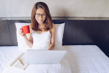 Portrait of Asian woman sitting on the bed and working on laptop with a red cup of coffee. Conceptual of woman lifestyle during working at home.