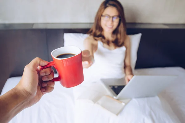 Portrait of someone giving a cup of coffee to Asian woman while her working on the bed. Conceptual of couple lifestyle.