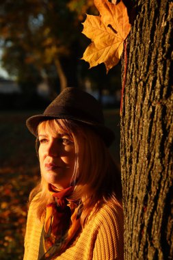 Renkli turuncu sonbahar parkındaki güzel sarışın kadının yakın plan portresi. Şapkalı ve ağaç gövdesinin yanında örgü ören şık bir kadın. Merhaba sonbahar konsepti. Sarı akçaağaç yaprağı. Aşk doğası.